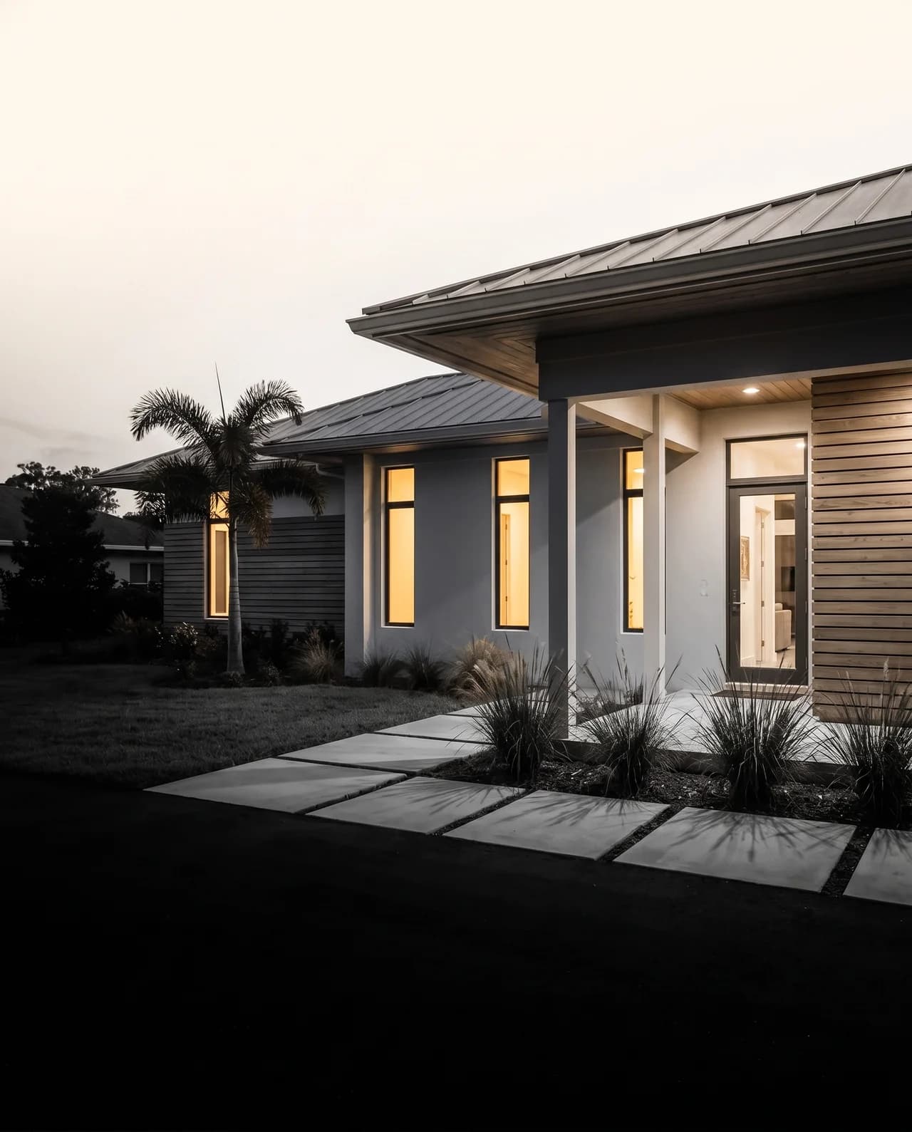 A modern Florida home's entrance glowing at dusk, palms and native grasses along the walk